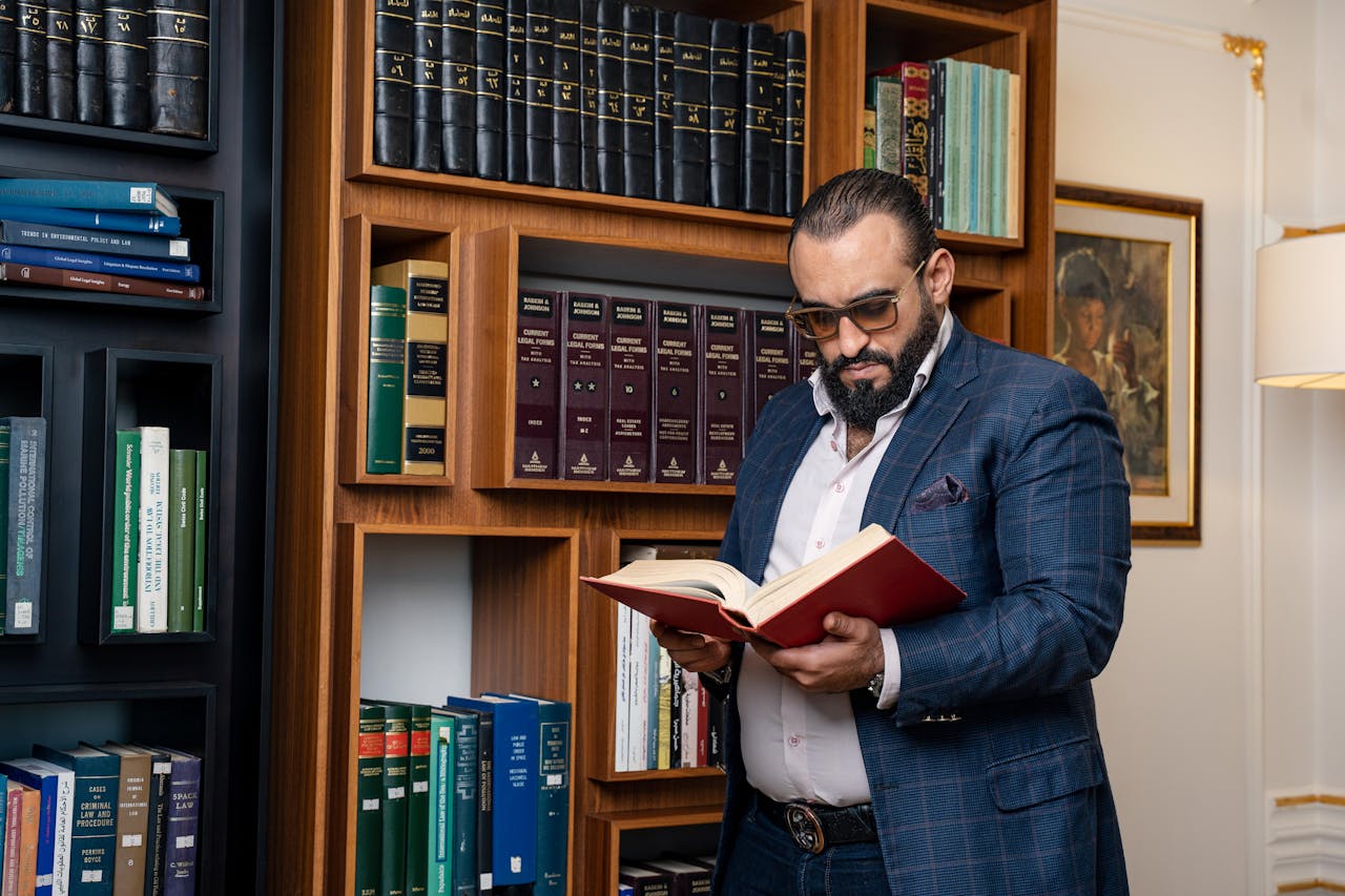Man with beard and glasses reading a book in a sophisticated library setting.